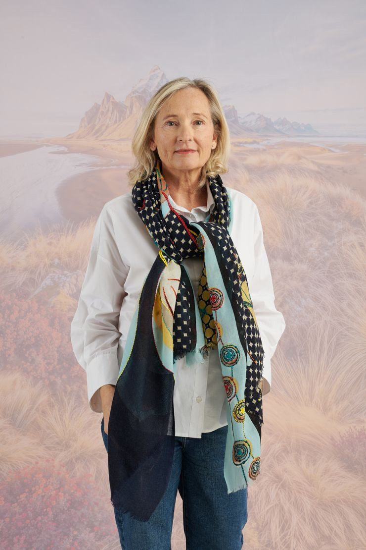 A woman with long blonde hair smiles at the camera in a scenic setting, wearing a white shirt, plaid pants, and the STORIATIPIC - ASTER WOOL SCARF by STORIATIPIC. Mountains, grass, and water are visible in the background.
