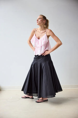 A woman with blonde hair tied back stands barefoot on a light floor, wearing a light pink spaghetti strap top and the HOPE FOR FLOWERS - CIRCLE SKIRT IN BLACK, made from GOTS Certified organic cotton, facing away against a plain wall.
