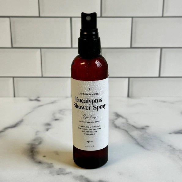 A hand holds a brown spray bottle labeled “SPA DAY EUCALYPTUS SHOWER SPRAY” by GIFTEN MARKET—an uplifting eucalyptus aromatherapy mist—set against a black showerhead and white marble shower wall, ideal for transforming your shower into a spa-like escape at home.