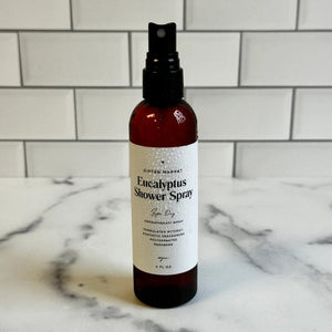 A hand holds a brown spray bottle labeled “SPA DAY EUCALYPTUS SHOWER SPRAY” by GIFTEN MARKET—an uplifting eucalyptus aromatherapy mist—set against a black showerhead and white marble shower wall, ideal for transforming your shower into a spa-like escape at home.