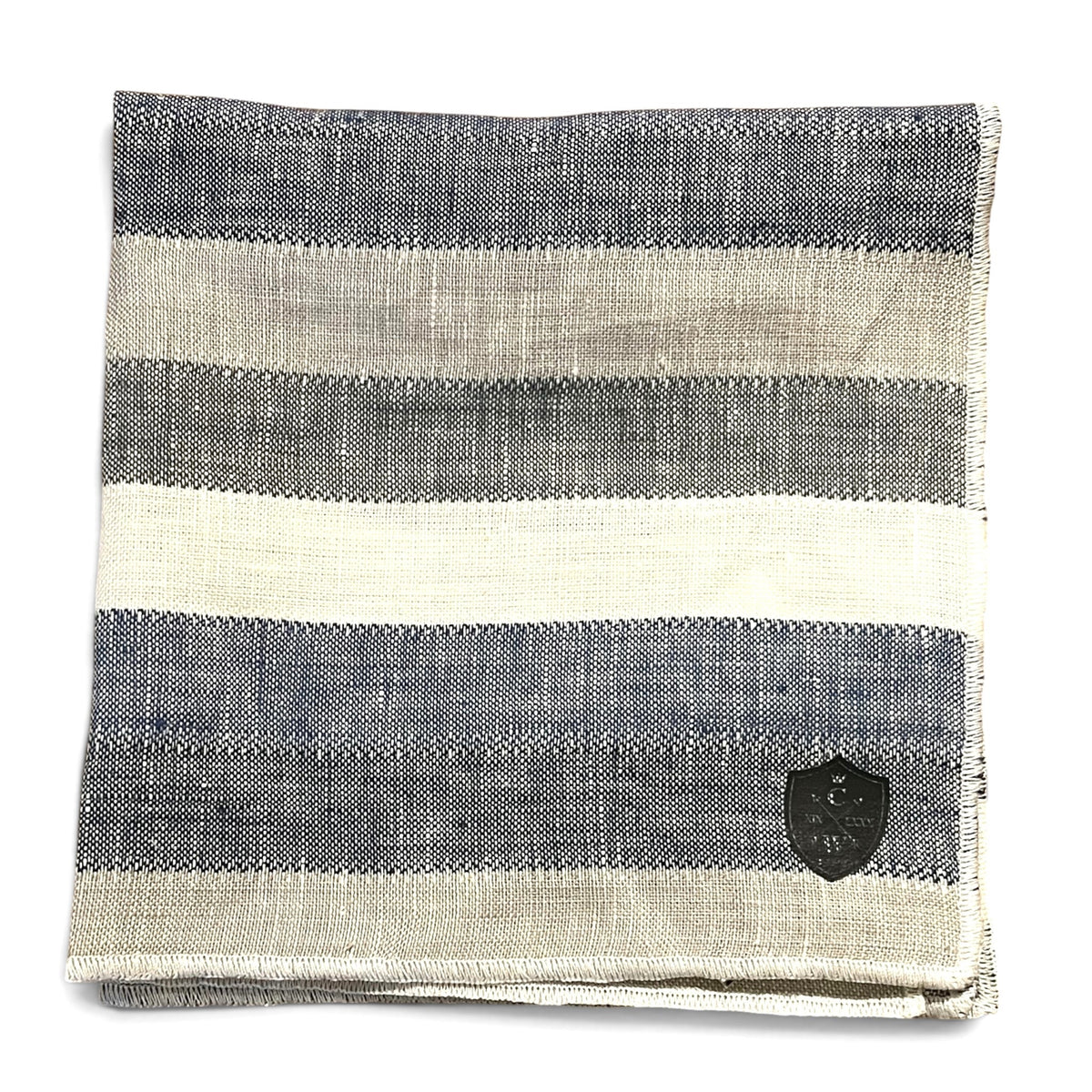The CLIFTON WILSON BLUE STRIPE LINEN POCKET SQUARE features wide faded stripes in grey, beige, and off-white with a dark shield-shaped patch in one corner; proudly made in the USA.