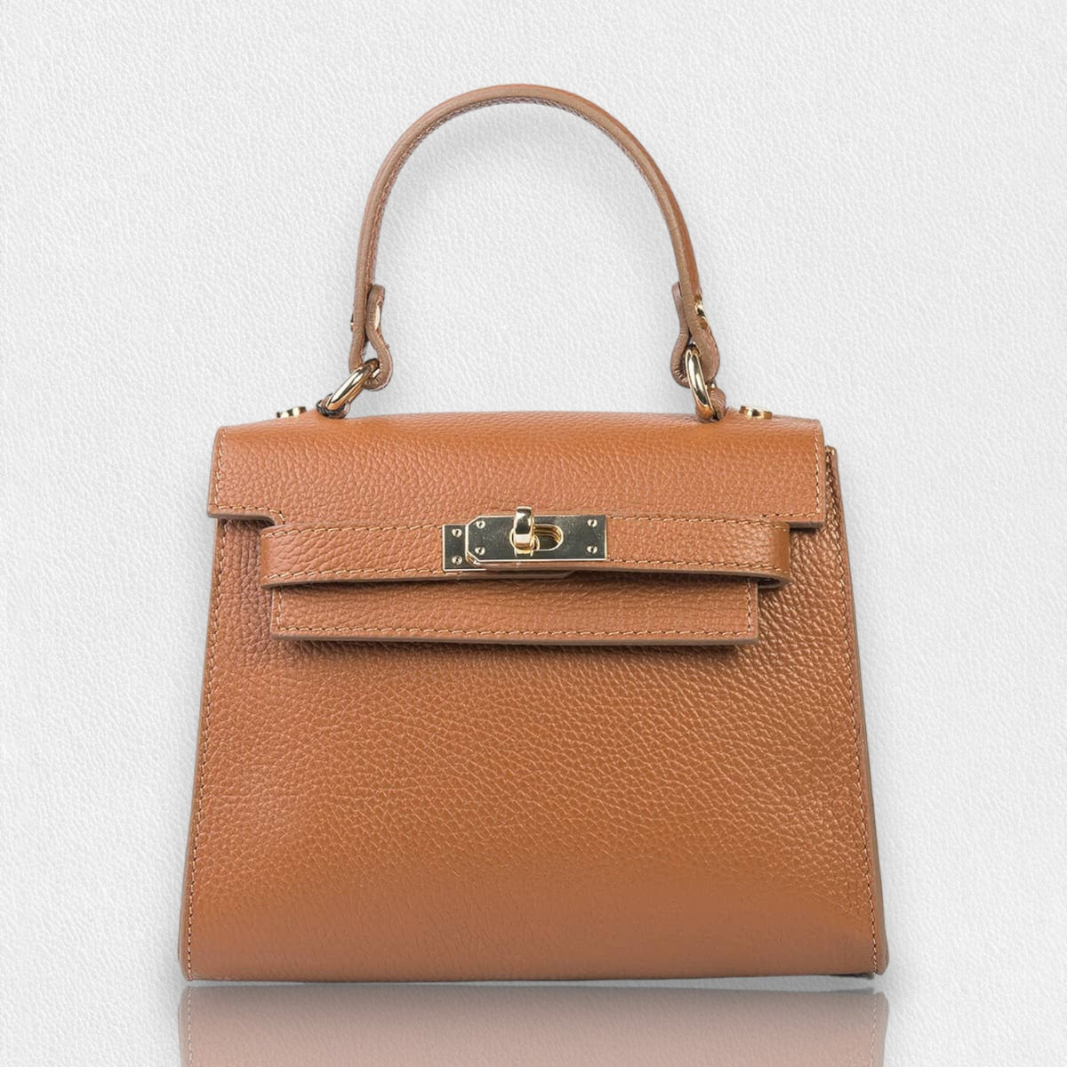 The LE BORSETTE LEATHER SATCHEL BAG is a small, structured satchel-shaped handbag in light pink, featuring a gold clasp at the front. It boasts a single top handle, an adjustable shoulder strap, and two gold-tone hardware pieces on either side of the handle. The textured leather adds to its luxurious and elegant appearance against the plain white background.
