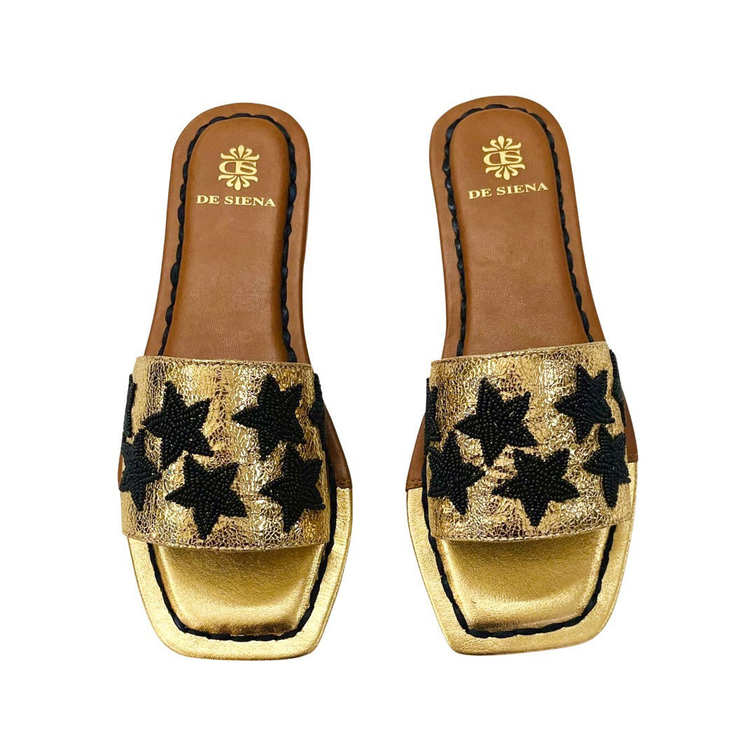 Introducing the DA SIENA – BEADED STARS SLIDES by DE SIENA. These open-toe leather slides feature brown soles and exquisite gold detailing. The straps are elegantly adorned with black beaded stars, while the cushioned insole in light brown showcases the DE SIENA logo and name. The sandals boast a chic flat, rectangular design.