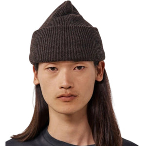 A person with long dark hair wears the HARTFORD - SEAMLESS WOOL BEANIE IN BROWN and a dark shirt, looking directly at the camera against a white background. The 100% wool beanie from HARTFORD adds both warmth and style to their look.