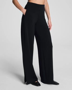 A person stands with one hand in their pocket, wearing SPANX AirEssentials Zero Waistband Wide Leg Pant made from light-as-air fabric and pointed black shoes against a plain light background.