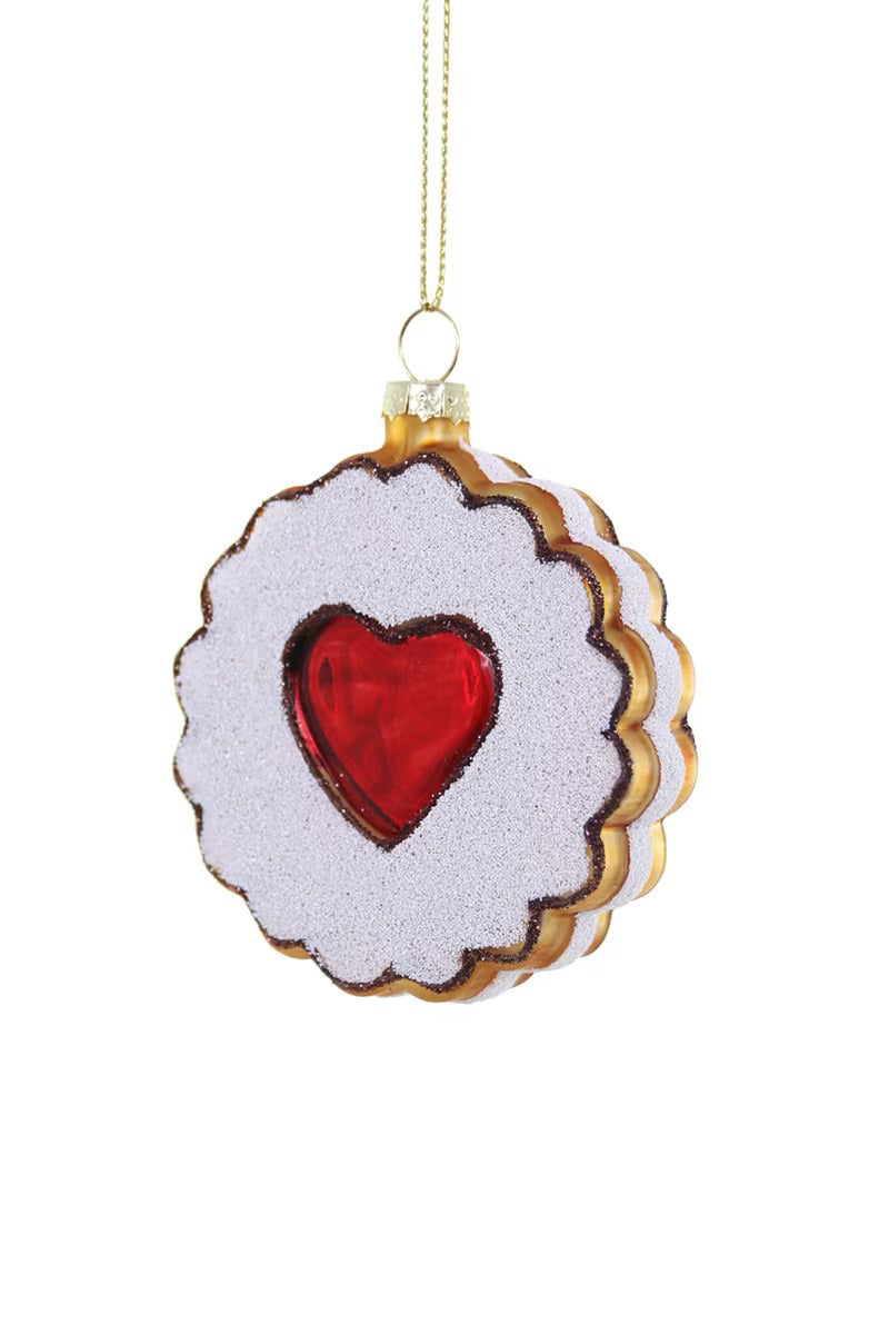 The LINZER HEART COOKIE ORNAMENT by CODY FOSTER is a glass Christmas tree ornament shaped like a round sandwich cookie with scalloped edges, white icing, and a red heart center, hanging by a gold string.