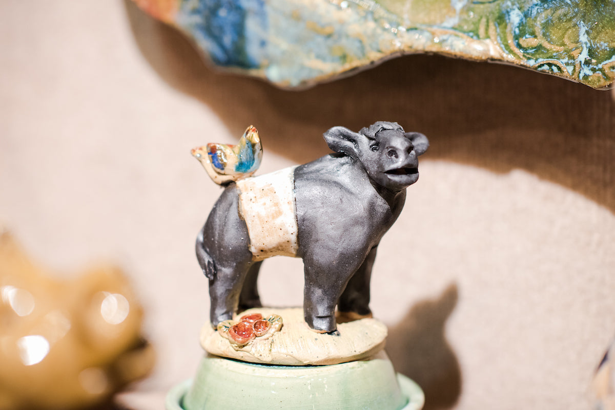 A small ceramic sculpture of a black and white cow with a bird perched on its back, standing on a green base with a small rose decoration. The background is softly blurred.