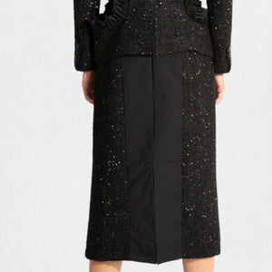 A person is wearing a stylish, textured black MEIMEIJ Lurex Tweed suit, featuring a tailored jacket with fringe details and the MEIMEIJ - LUREX TWEED SKIRT. Underneath the jacket is a white T-shirt with bold black text. The outfit is paired with black socks and black shoes.