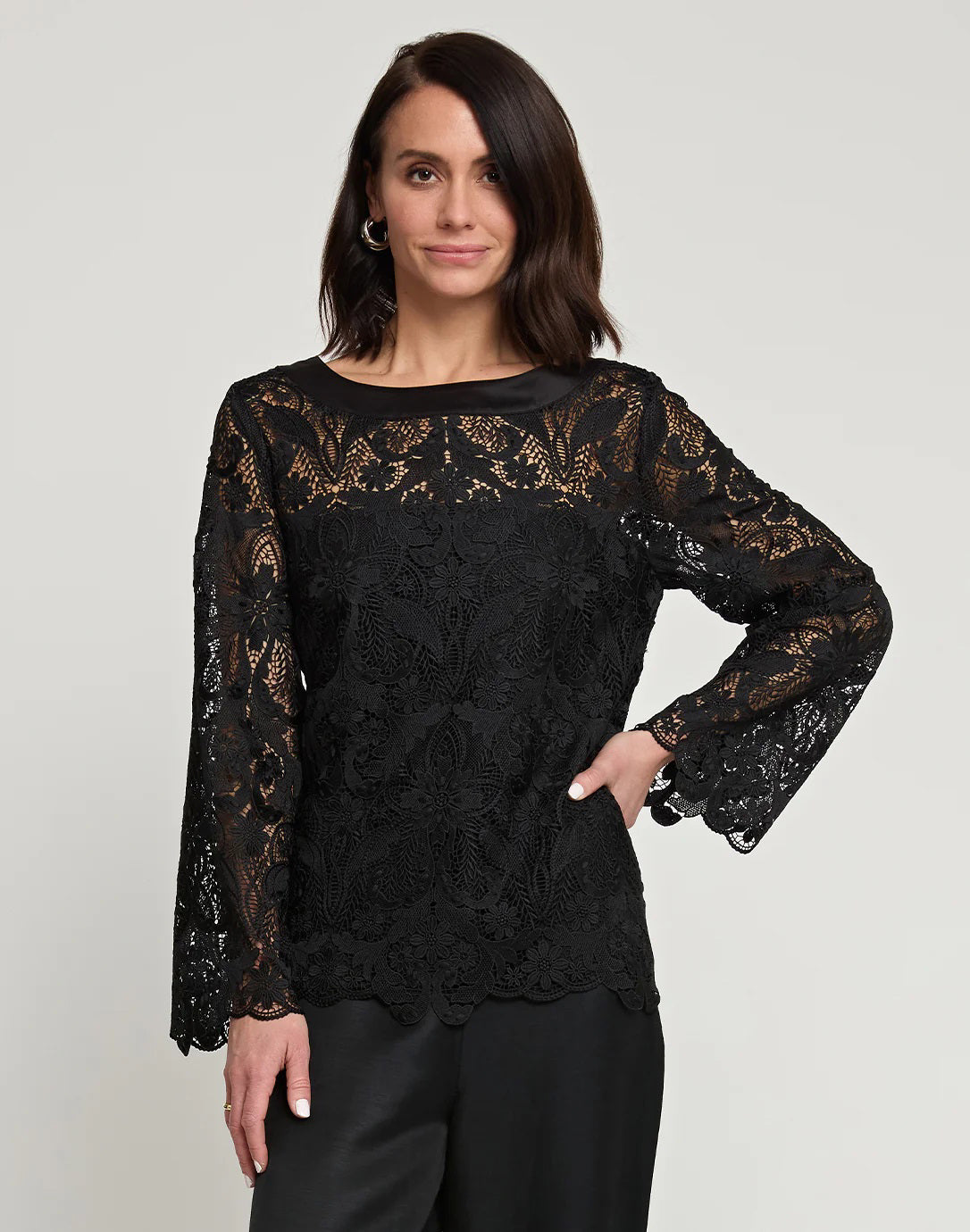 A woman with shoulder-length dark hair is wearing the HINSON WU - BLAKELY LACE TOP, a long-sleeve black lace top that features flared sleeves, paired with black pants. She stands against a plain light gray background, neutrally expressing herself with one hand on her hip.