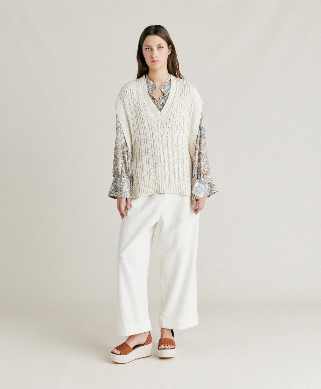 A woman with straight brown hair stands against a plain background. She is wearing the MOMONI - ARENE KNIT VEST from MOMONI, an oversized, cream-colored cable knit vest made from cotton yarn, over a patterned, long-sleeve button-up shirt and paired with white pants. Her hands are in her pockets, and she has a neutral expression.