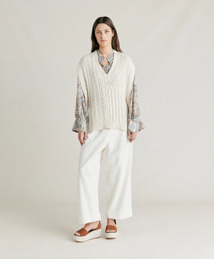 A woman with straight brown hair stands against a plain background. She is wearing the MOMONI - ARENE KNIT VEST from MOMONI, an oversized, cream-colored cable knit vest made from cotton yarn, over a patterned, long-sleeve button-up shirt and paired with white pants. Her hands are in her pockets, and she has a neutral expression.