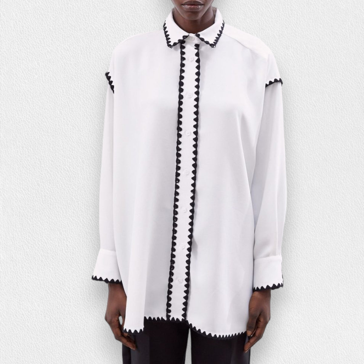 A person wears the MEIMEIJ - TUNIC LENGTH BLOUSE by MEIMEIJ, a loose-fit 100% viscose top with black edge details on the collar, placket, cuffs, and hem, paired with black pants against a light textured background.