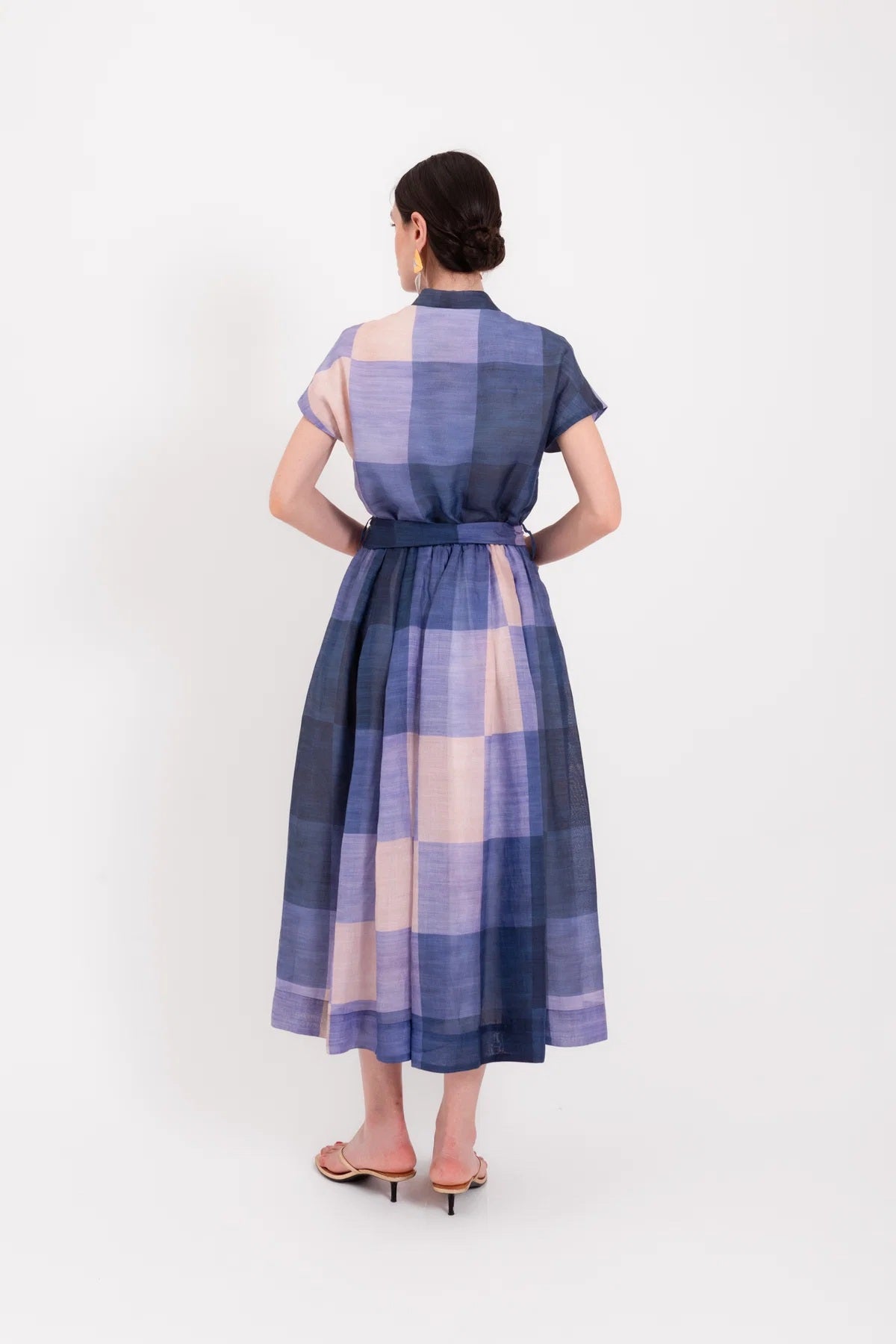 A woman wearing the LAVI - BELLELL Dress in Plaid by LAVI, a short-sleeve, collared shirt dress with a blue and beige plaid pattern and a tied waist, accessorized with gold earrings, bracelets, and open-toed heels, stands against a plain white background.