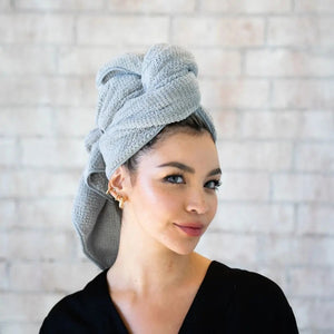 A person with short dark hair, wearing a white shirt, wraps their hair in the VOLO BEAUTY - HERO HAIR TOWEL LUXE and adjusts it with both hands in a bright, airy space filled with soft natural light.
