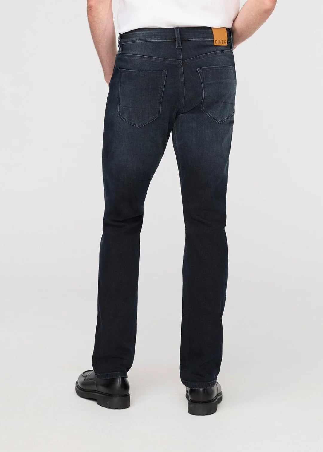 A man stands against a plain background wearing a black jacket, white t-shirt, DU/ER - PERFORMANCE DENIM+ SLIM STRIAGHT PANTS IN CARBON STONE, and black shoes, with one hand in his pocket and a neutral expression.