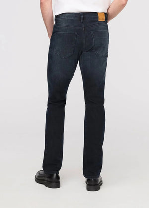 A man stands against a plain background wearing a black jacket, white t-shirt, DU/ER - PERFORMANCE DENIM+ SLIM STRIAGHT PANTS IN CARBON STONE, and black shoes, with one hand in his pocket and a neutral expression.