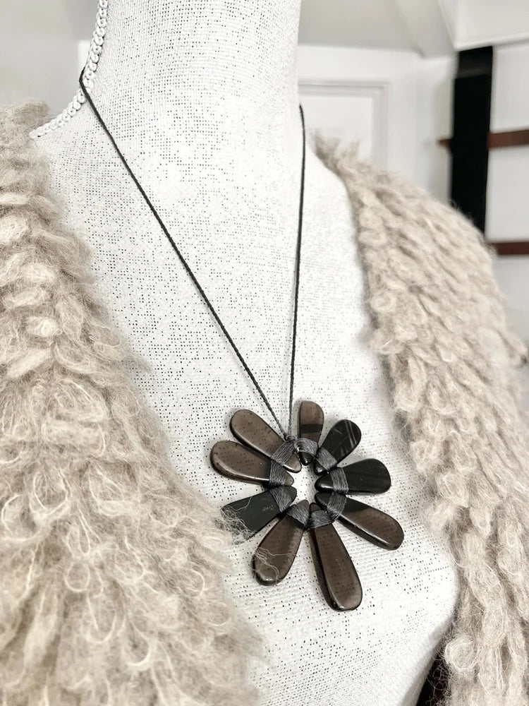 A mannequin wears a shaggy beige jacket paired with the ANN LIGHTFOOT Nine Petal Smokey Quartz Flower Necklace, which features a modern black cord and a bold, dark metallic floral pendant.