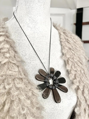 A mannequin wears a shaggy beige jacket paired with the ANN LIGHTFOOT Nine Petal Smokey Quartz Flower Necklace, which features a modern black cord and a bold, dark metallic floral pendant.