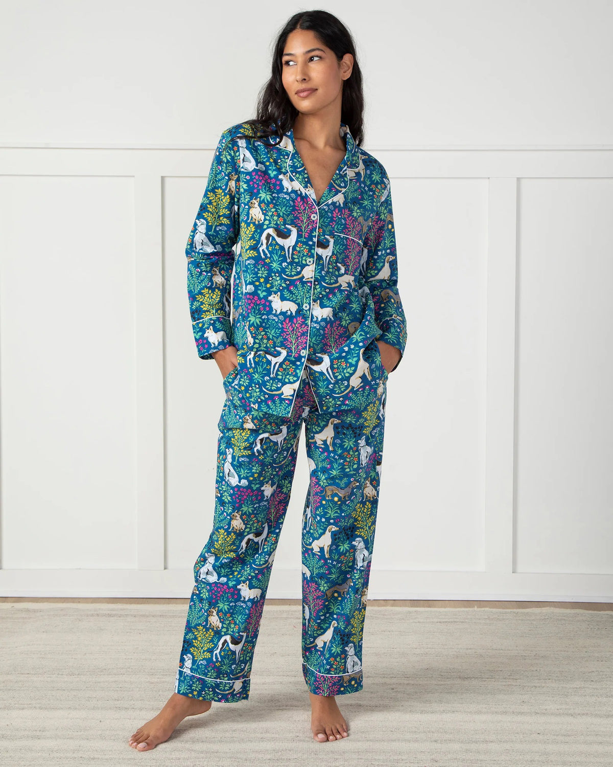 A woman in PRINTFRESH&#39;s colorful &quot;Must Love Dogs&quot; organic cotton pajama set stands barefoot on a light rug against a white paneled wall. Her dark hair falls freely as she casually places her hands in her pockets, facing slightly to the side.