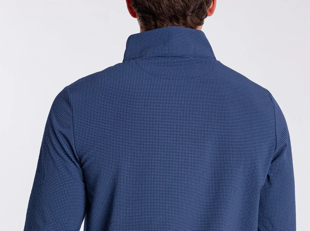 A man in the CRIQUET Seersucker Performance Pullover in Navy and light pants smiles with his hand by his side; this lightweight layer from CRIQUET offers UPF 50 protection against a simple background.