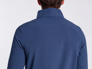 A man in the CRIQUET Seersucker Performance Pullover in Navy and light pants smiles with his hand by his side; this lightweight layer from CRIQUET offers UPF 50 protection against a simple background.