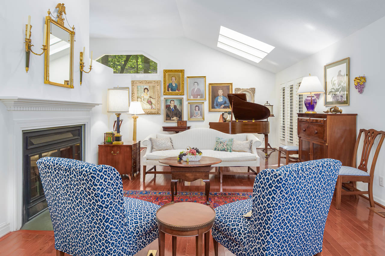 A bright, elegant living room with blue patterned chairs, a white sofa, a grand piano, framed portraits on the walls, a fireplace, wooden furniture, and natural light from skylights.