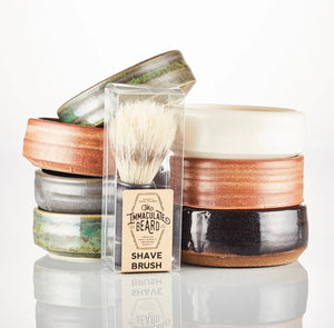 THE IMMACULATE BEARD - SHAVE BRUSH by THE IMMACULATE BEARD, featuring a black handle and faux boar bristles, stands upright on a wooden tray with a green plant and white tiled wall blurred in the background.