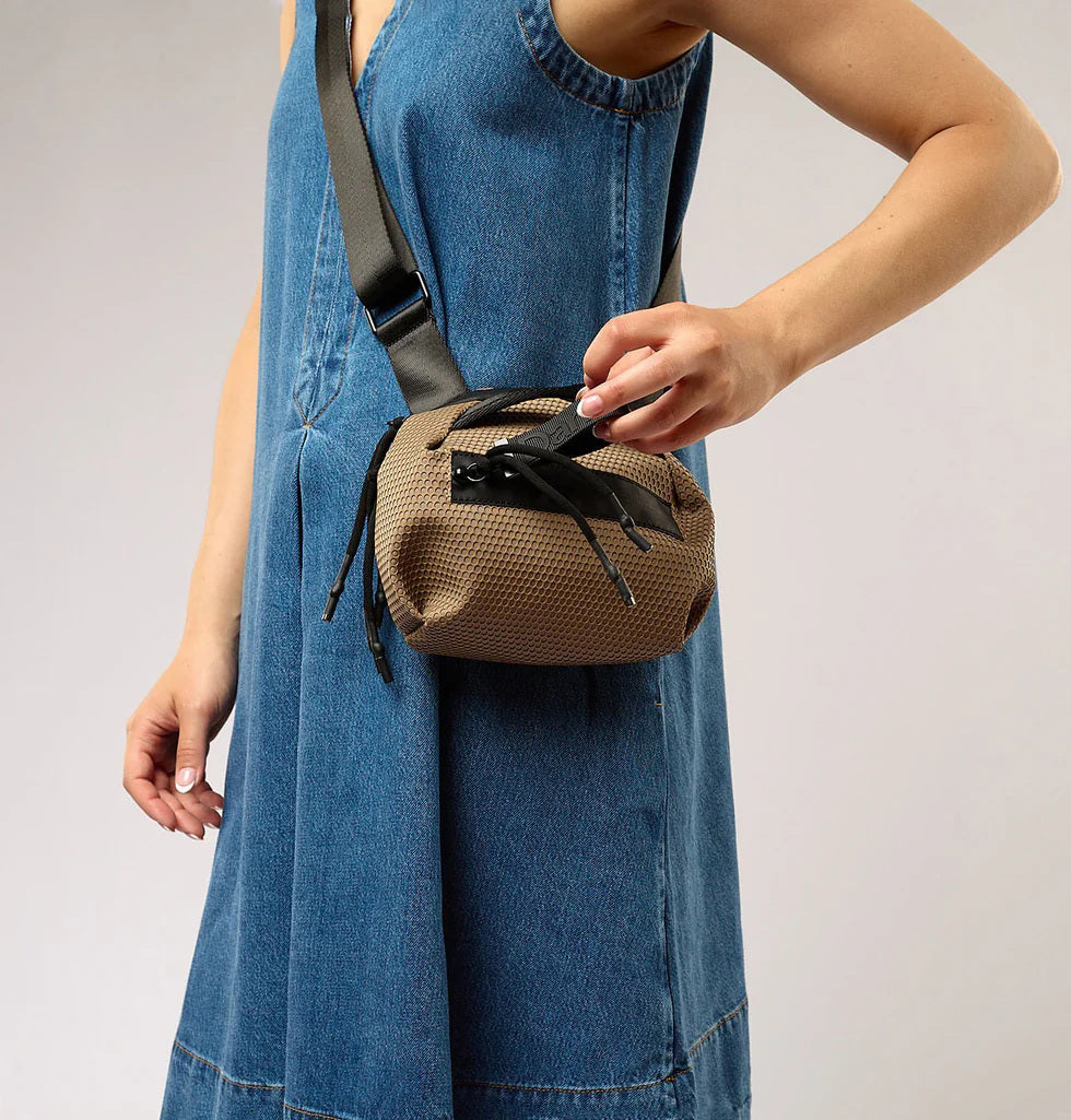 The DANIELLA LEHAVI BONNIE MINI TRAVEL CROSSBODY, a small brown vegan bag with black accents and strap, is displayed on a white pedestal against a plain beige background.