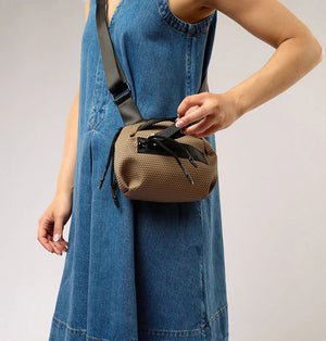 The DANIELLA LEHAVI BONNIE MINI TRAVEL CROSSBODY, a small brown vegan bag with black accents and strap, is displayed on a white pedestal against a plain beige background.