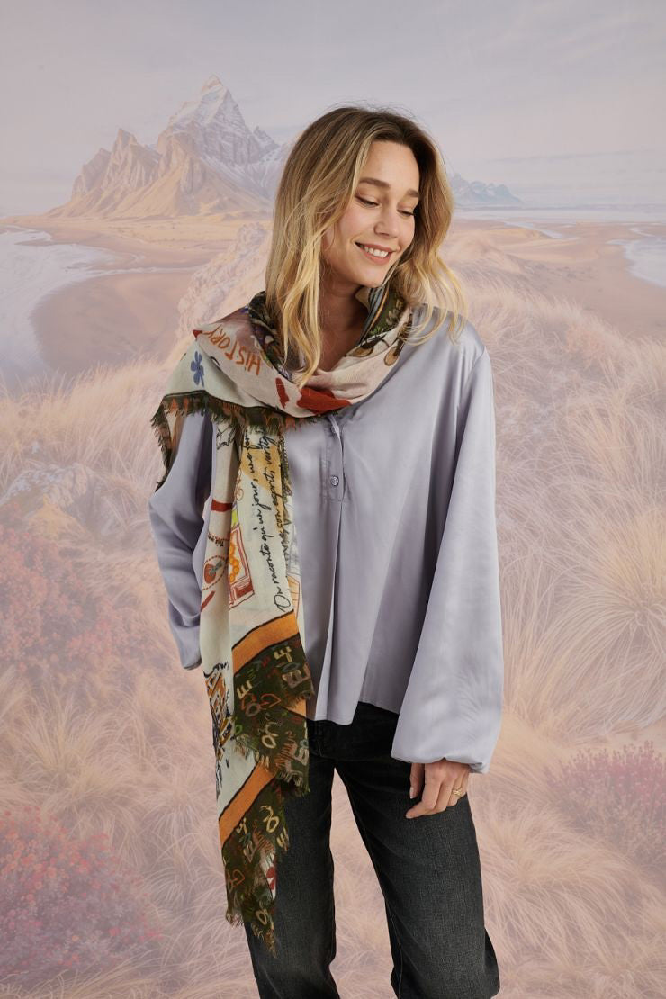A woman with wavy blonde hair smiles while looking down. She wears a loose, light lavender blouse, dark pants, and the STORIATIPIC EUGENIE WOOL SCARF. Behind her is a scenic landscape of mountains, grass, and water.