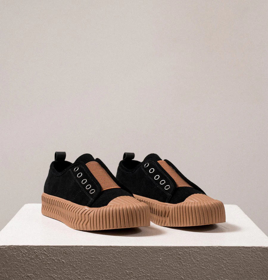 Introducing the DANIELLA LEHAVI - OSAKA SNEAKER by DANIELLA LEHAVI: Black sneakers featuring brown rubber soles and decorative brown straps across the top. They highlight a contemporary design with circular metal eyelets on the sides and are presented on a white platform against a simple, light-colored background.