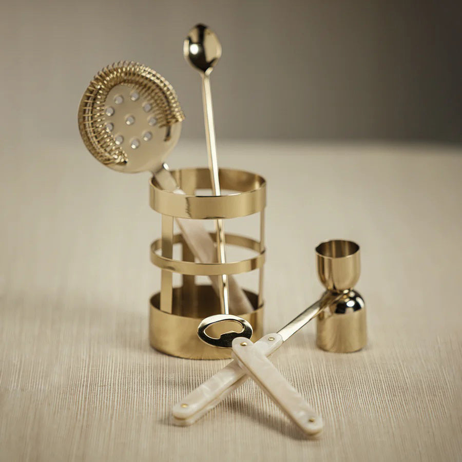 A set of elegant gold bar tools with white handles, including a strainer, spoon, jigger, and bottle opener, arranged neatly in and around a matching gold holder on a beige surface.