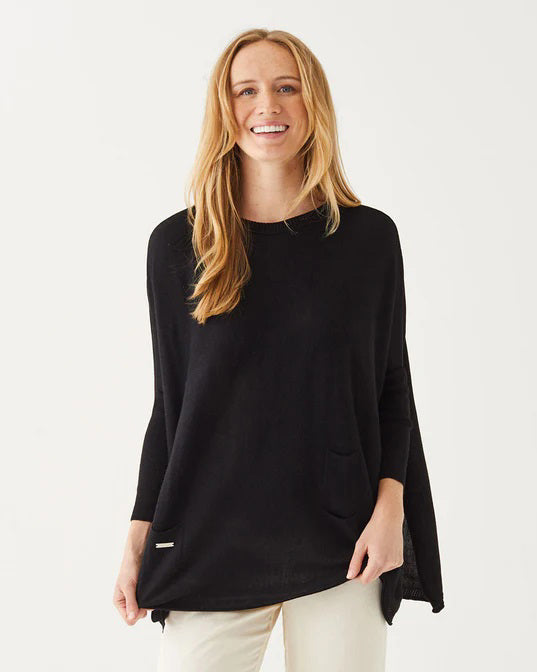 A smiling woman with long blonde hair wears the MERSEA Catalina Crewneck Sweater in black, featuring signature split sides, paired with light pants and standing against a plain white background.