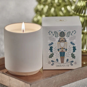A plain NUTCRACKER SIBERIAN FIR CANDLE in a matte vessel sits next to its square box featuring a nutcracker illustration and holly motifs.