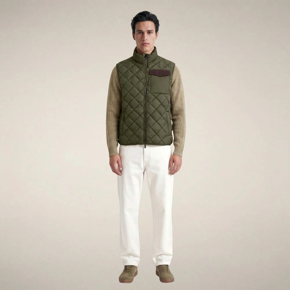 A person stands facing forward, wearing the SAVE THE DUCK - FABRICE Puffer Vest in Snake Green with Plumtech® padding over a beige sweater, paired with white pants and tan shoes, against a plain light background.