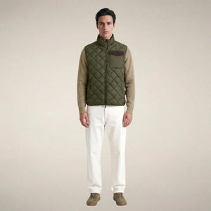 A person stands facing forward, wearing the SAVE THE DUCK - FABRICE Puffer Vest in Snake Green with Plumtech® padding over a beige sweater, paired with white pants and tan shoes, against a plain light background.