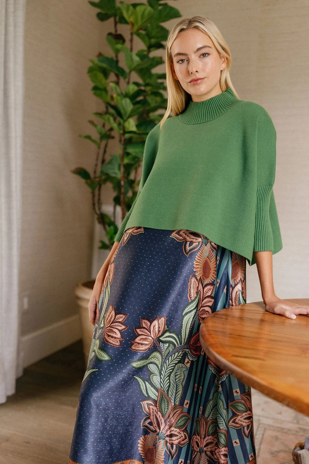 A woman stands indoors by a table, wearing the BRITON COURT SOPHIA Oversized One Size Sweater in Evergreen, paired with a long, floral-patterned skirt in earthy tones. A leafy potted plant is visible in the background.