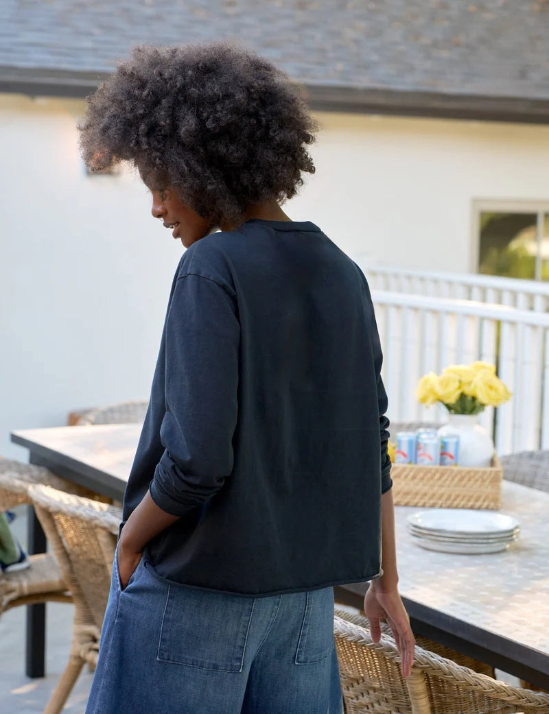 A woman with naturally curly hair, wearing the FRANK & EILEEN QUINN Long Sleeve Crewneck Tee in British Royal Navy and relaxed blue jeans, stands by an outdoor table with plates, drinks, and flowers, smiling softly. Made in California.