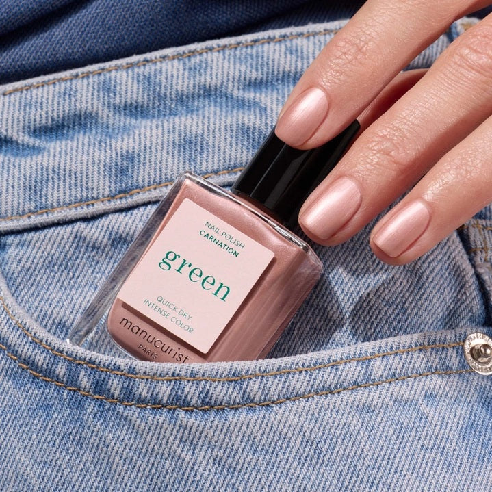 A bottle of MANUCURIST INC’s GREEN™ Nail Polish in the shade “Carnation,” featuring a black cap and pale pink color, with a label that reads “Quick Dry Intense Color.”.