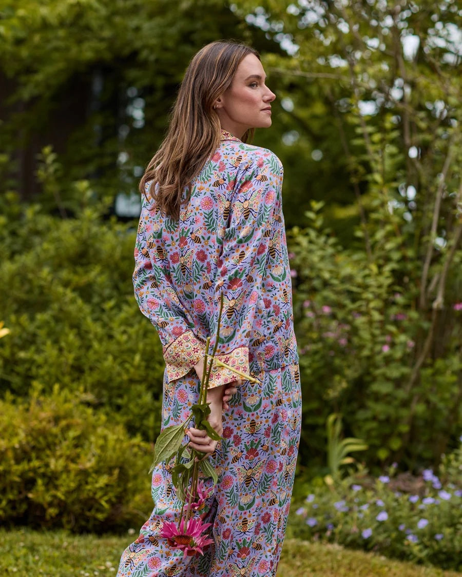 A person wearing the PRINTFRESH - BEE TAPESTRY ECO SATIN SOHO SET IN LAVENDER, featuring vintage-inspired bee and floral vine prints, holding a flower behind their back in a garden.