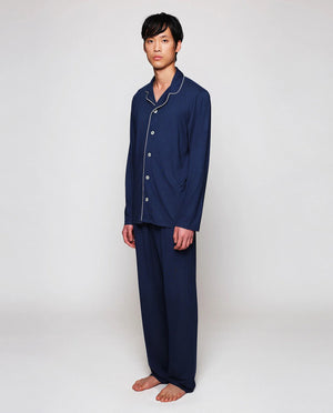 A person with short dark hair stands barefoot, facing sideways and looking back at the camera, wearing MIRTO 1956 Navy Modal Pajamas by MIRTO 1956 against a plain light background.