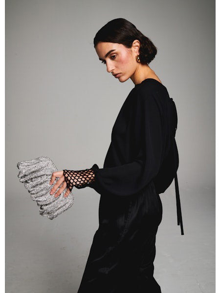 A woman with short dark hair faces away, wearing the MILVA MI - CROCHET CUFF BLOUSE by MILVA, black pants, and pointed black shoes. She holds a silver textured clutch against a plain gray background.