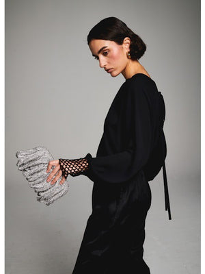 A woman with short dark hair faces away, wearing the MILVA MI - CROCHET CUFF BLOUSE by MILVA, black pants, and pointed black shoes. She holds a silver textured clutch against a plain gray background.