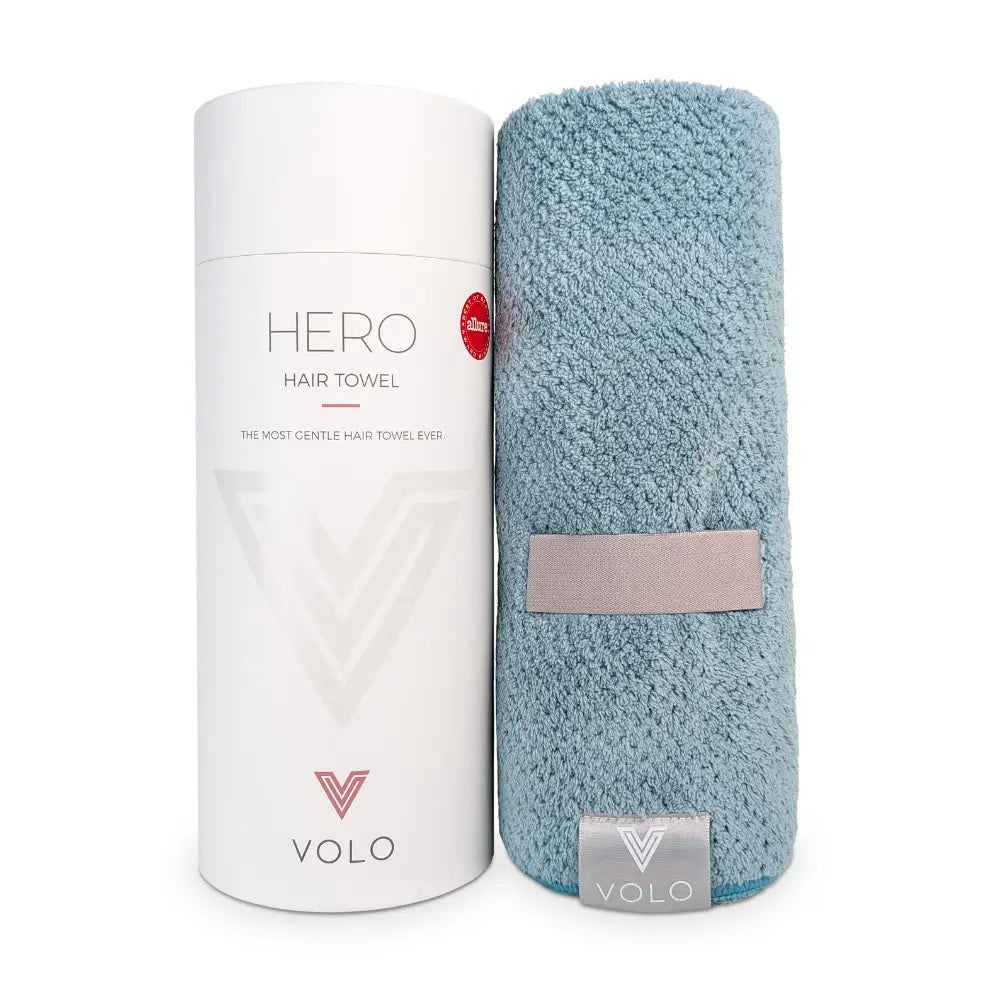 A person with short dark hair, wearing a white shirt, wraps their hair in the VOLO BEAUTY - HERO HAIR TOWEL LUXE and adjusts it with both hands in a bright, airy space filled with soft natural light.