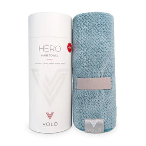 A person with short dark hair, wearing a white shirt, wraps their hair in the VOLO BEAUTY - HERO HAIR TOWEL LUXE and adjusts it with both hands in a bright, airy space filled with soft natural light.