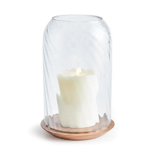 The NAPA HOME AND GARDEN - NIRA HURRICANE, LARGE features a white sculpted candle with a gold accent beneath ribbed blown glass on an acacia wood base, creating a striking decorative accent against a white background.