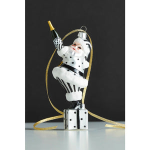 A CHAMPAGNE POPPING SANTA ORNAMENT by RAZ Imports is a resin Santa Claus wearing a silver and black outfit, holding a champagne bottle in one hand and standing on a gift box.