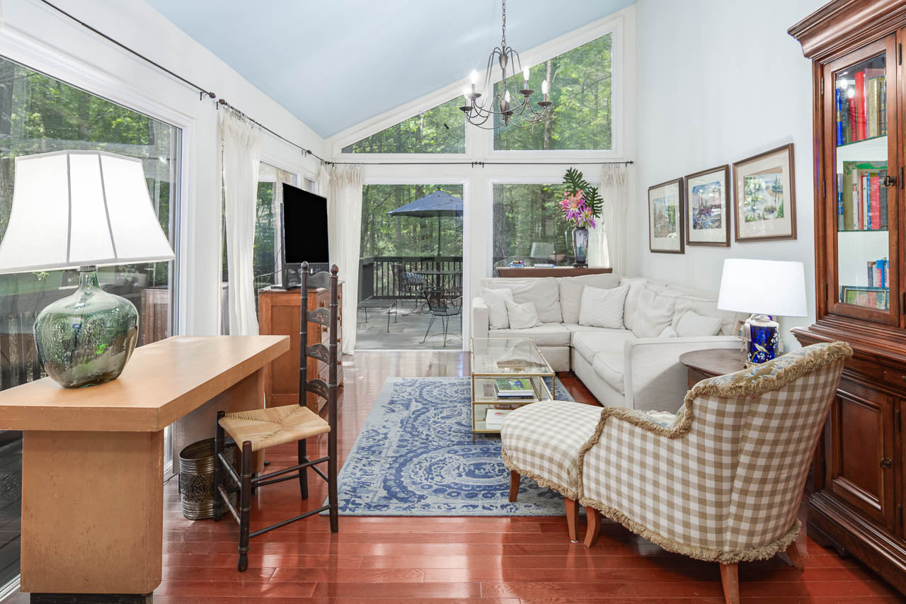 A bright living room with large windows, white walls, wood floors, a white sectional sofa, patterned armchair, wooden desk with a lamp, and a blue rug, overlooking a deck and green trees outside.