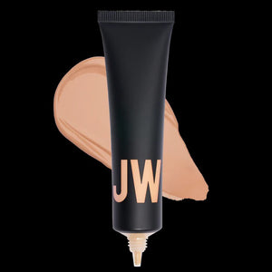 A close-up of a person with natural curly hair, adorned in gold hoop earrings and a white top. They wear a neutral facial expression while looking directly at the camera. The softly lit, plain background enhances their glowing skin as if touched by JASON WU - TINTED MOISTURIZER MEETS CC CREAM from JASON WU BEAUTY.