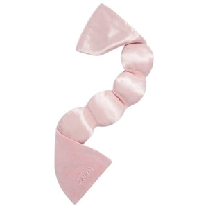 A woman in a gray bralette and shorts lies on white bedding, wearing the NODPOD Silk Weighted Sleep Eye Mask in Petal—a large pink satin silk mask shaped like a bow from NODPOD that covers her eyes for comfort and stress relief.